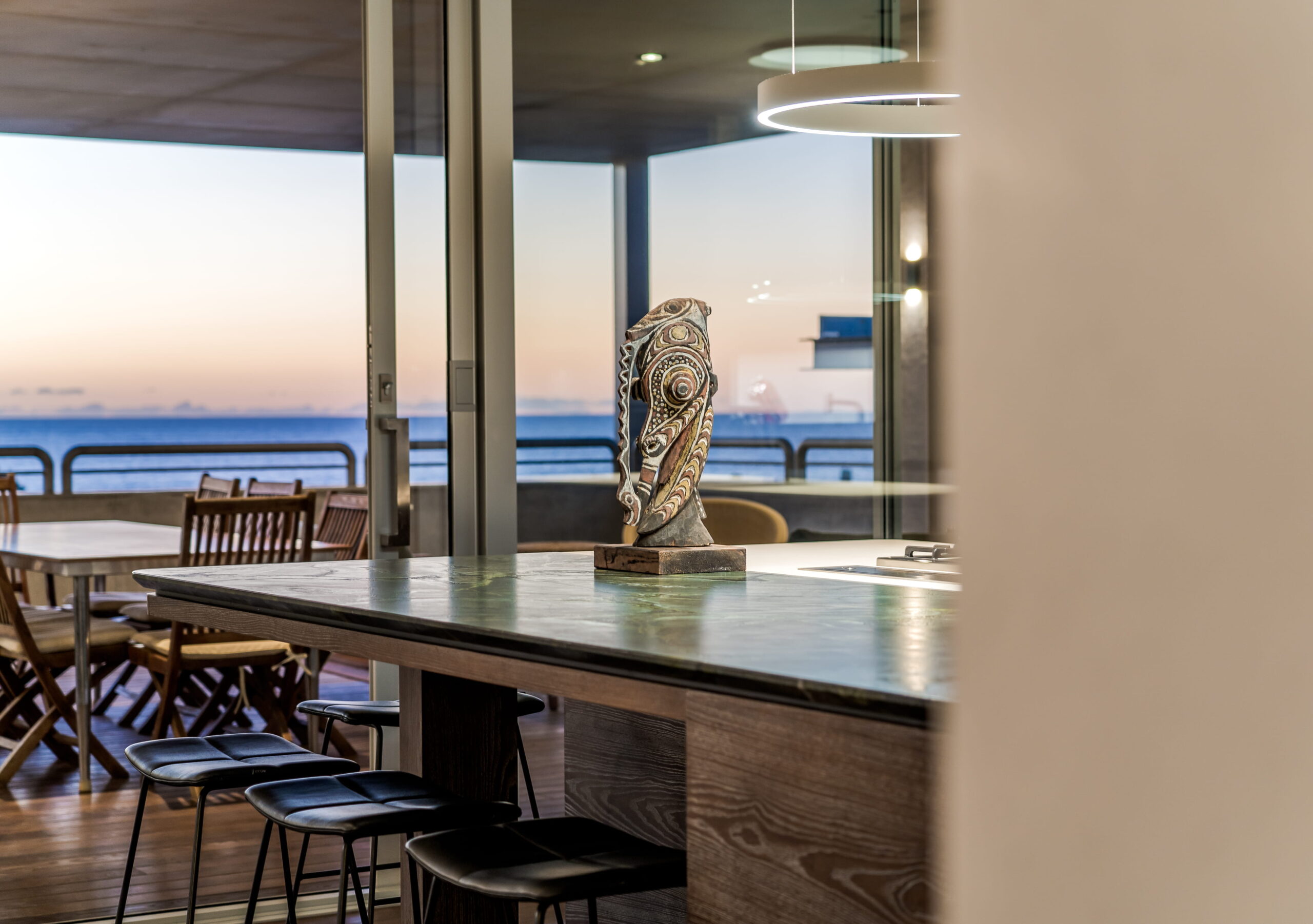 Interior View Looking Across a Kitchen Island Through Large Glass Sliding Doors to an Alfresco Area and Ocean Sunset.