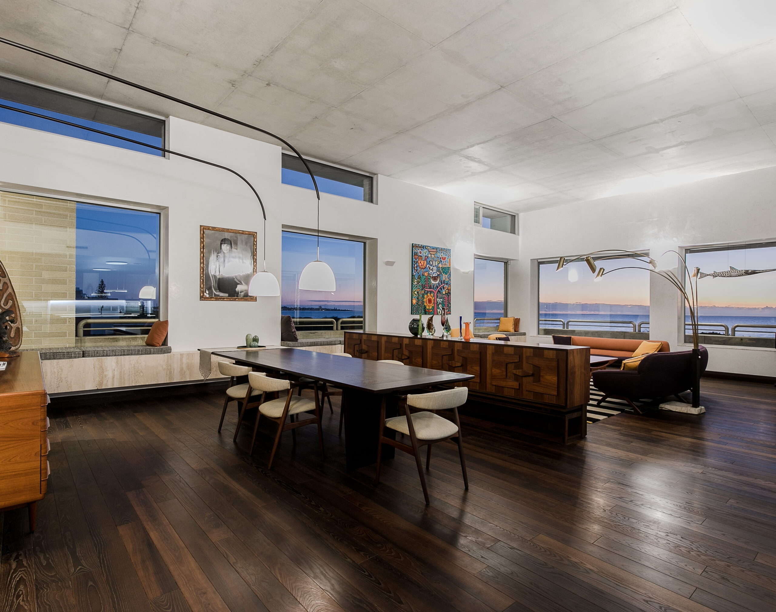 Modern Coastal Home Interior Featuring Dark Wood Floors and Large Windows Framing a Sunset View Over the Water.