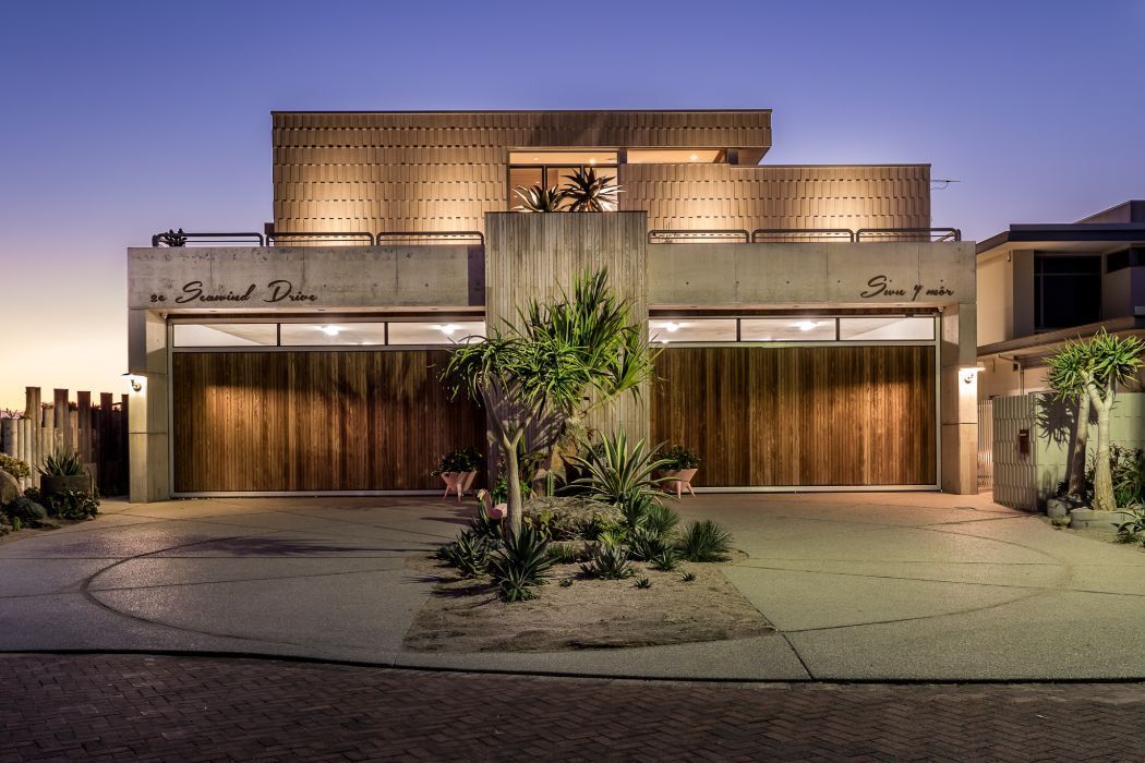 Modern luxury home facade with rooftop balcony, timber garage doors, and architectural concrete finishes.
